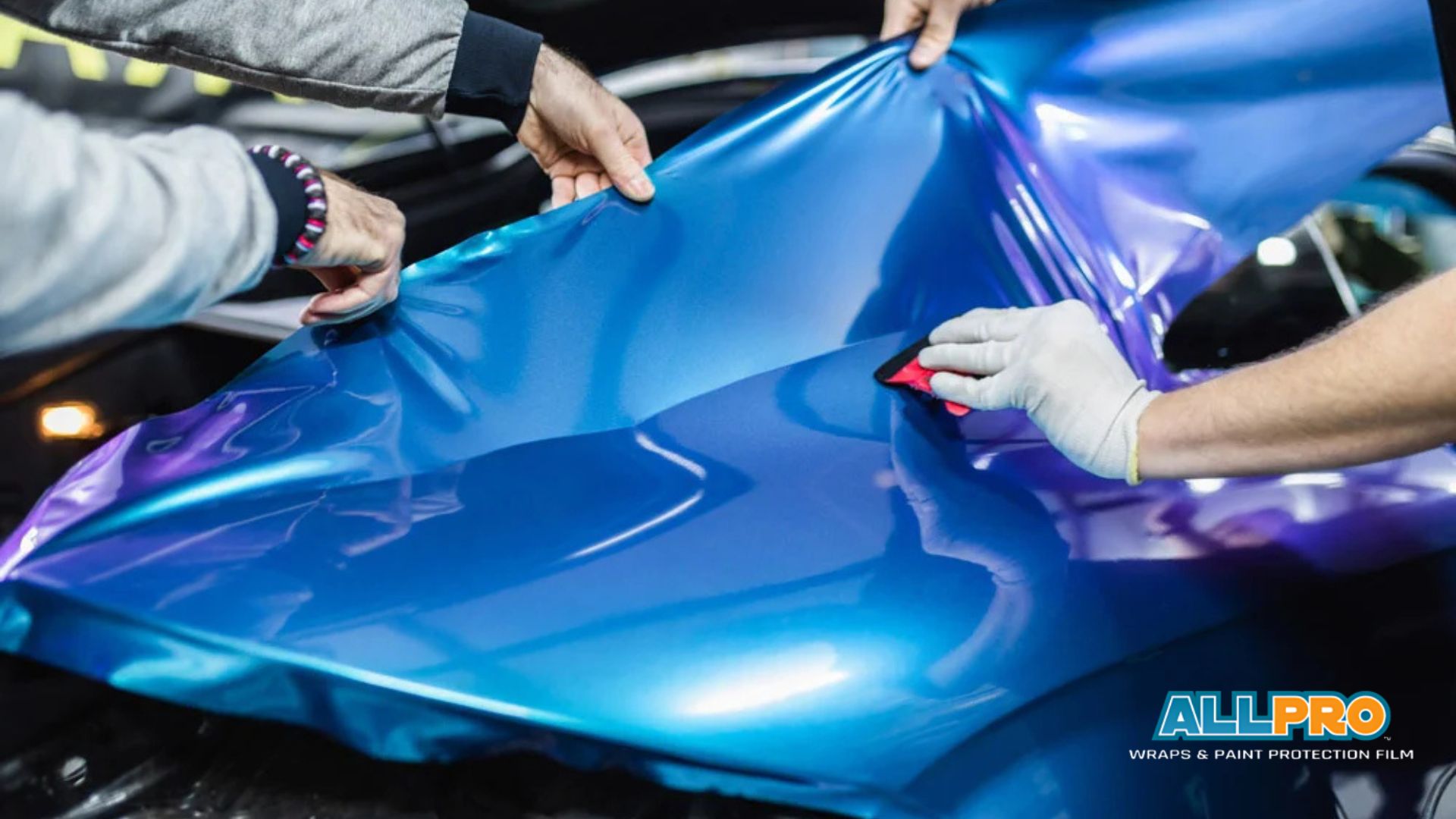 Technicians applying blue and purple vinyl wrap to a car hood using a squeegee to smooth the material and remove air bubbles during installation.