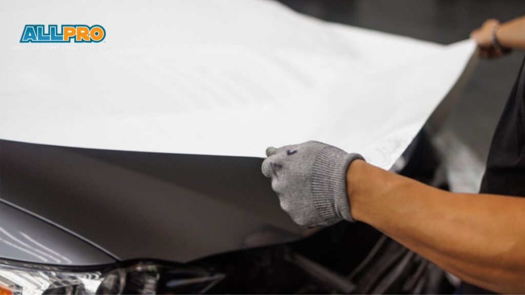 A technician wearing gray gloves stretches a large sheet of white vinyl wrap across the hood of a dark car during installation. The corner of the image shows the logo text "ALLPRO WRAPS & PAINT PROTECTION FILM" while the vinyl wrap is carefully positioned before being applied to the vehicle surface.