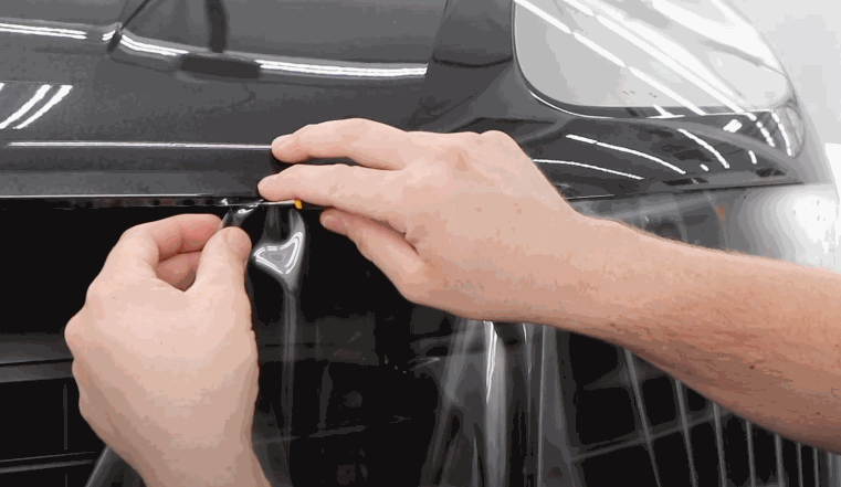 Hands carefully align and trim Paint Protection Film along the edge of a car front grille for a precise fit. The transparent film is positioned tightly against the painted surface to protect the front end from chips and road debris.