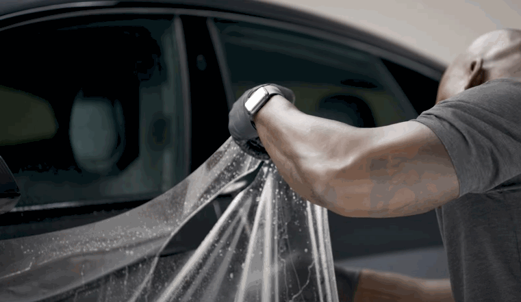 Technician stretches a clear Paint Protection Film across a car door while installing the protective layer on the vehicle exterior. Water droplets on the film help position the transparent sheet before it is smoothed onto the paint surface.
