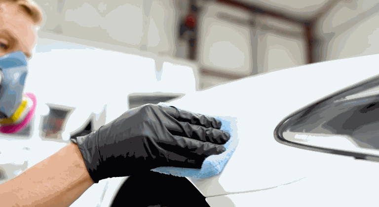 A technician wearing black gloves wipes down a white car panel with a blue microfiber cloth in a bright workshop. This prep step ensures the surface is clean and ready for ceramic coating application.
