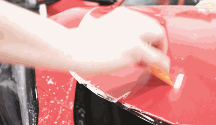 Close up of PPF being peeled back and adjusted on a red car surface with water droplets visible underneath the film. The scene shows paint protection film installation during alignment and repositioning.