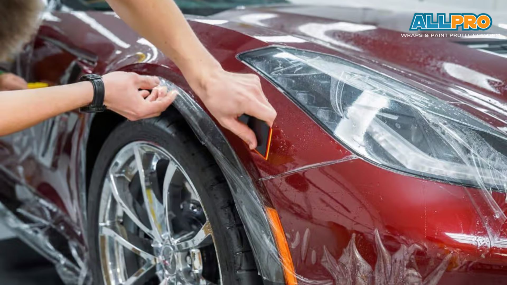 Technician applying PPF to a red car fender using a squeegee tool while stretching the film smoothly over the curved surface. The process shows paint protection film being installed to shield the paint from damage.