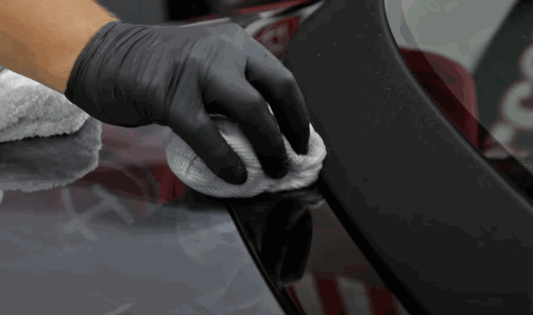 A gloved hand wipes down a black car panel with a microfiber cloth, removing residue and enhancing shine during car detailing. The surface appears smooth and reflective after cleaning.