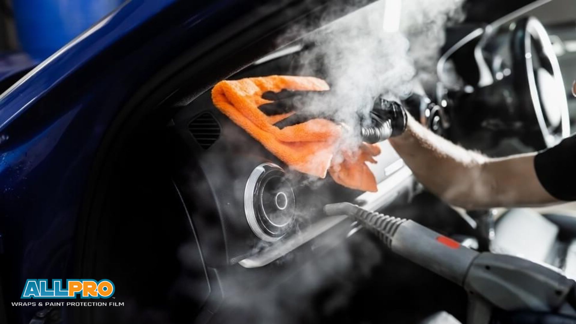 Steam cleaner and orange microfiber towel deep clean a car interior dashboard, lifting dirt and grime as part of a thorough car detailing service. Visible steam surrounds the control panel during the process.