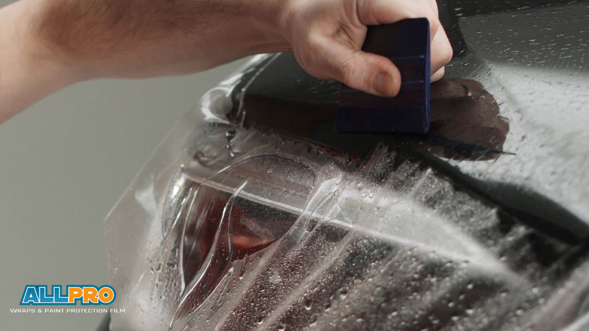 Hand uses a squeegee to smooth clear paint protection film over a car headlight with visible water droplets and the "ALL PRO Wraps & Paint Protection Film" logo in the corner.