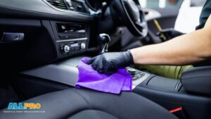 Gloved hand wiping a car center console with a purple microfiber cloth during car detailing.