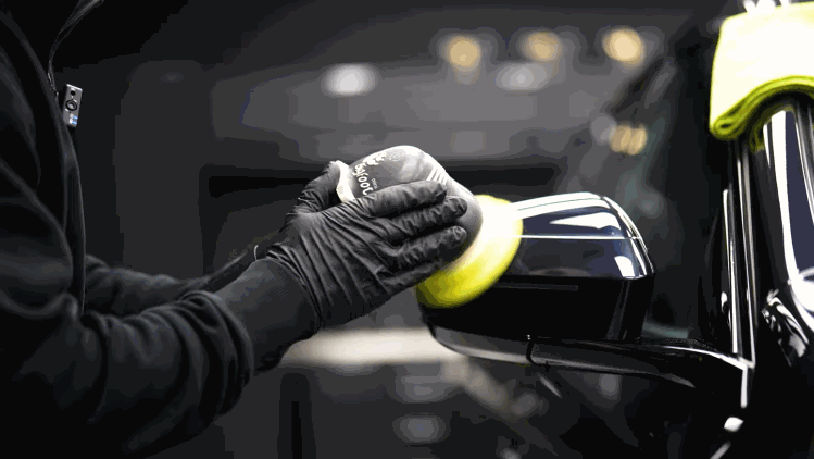 A technician in black gloves hand polishes the side mirror of a black vehicle before ceramic coating. The close up shows careful detailing around the exterior trim.