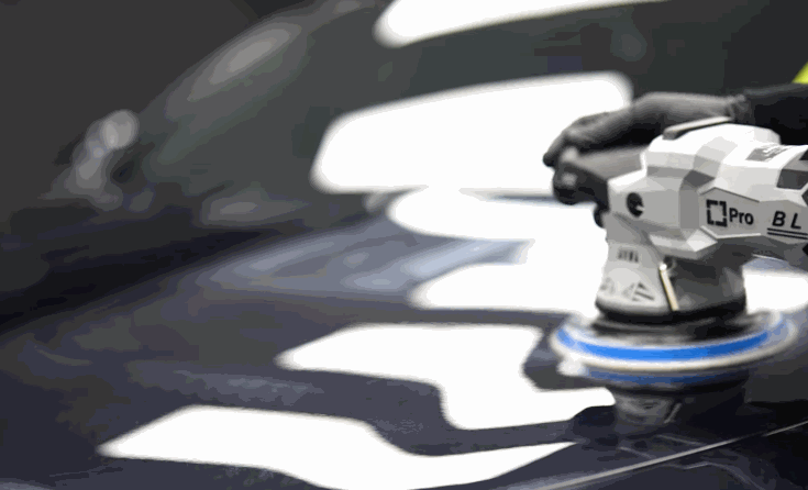 A machine polisher buffs the hood of a black vehicle to prepare the paint for ceramic coating. Bright workshop lights reflect across the glossy surface.