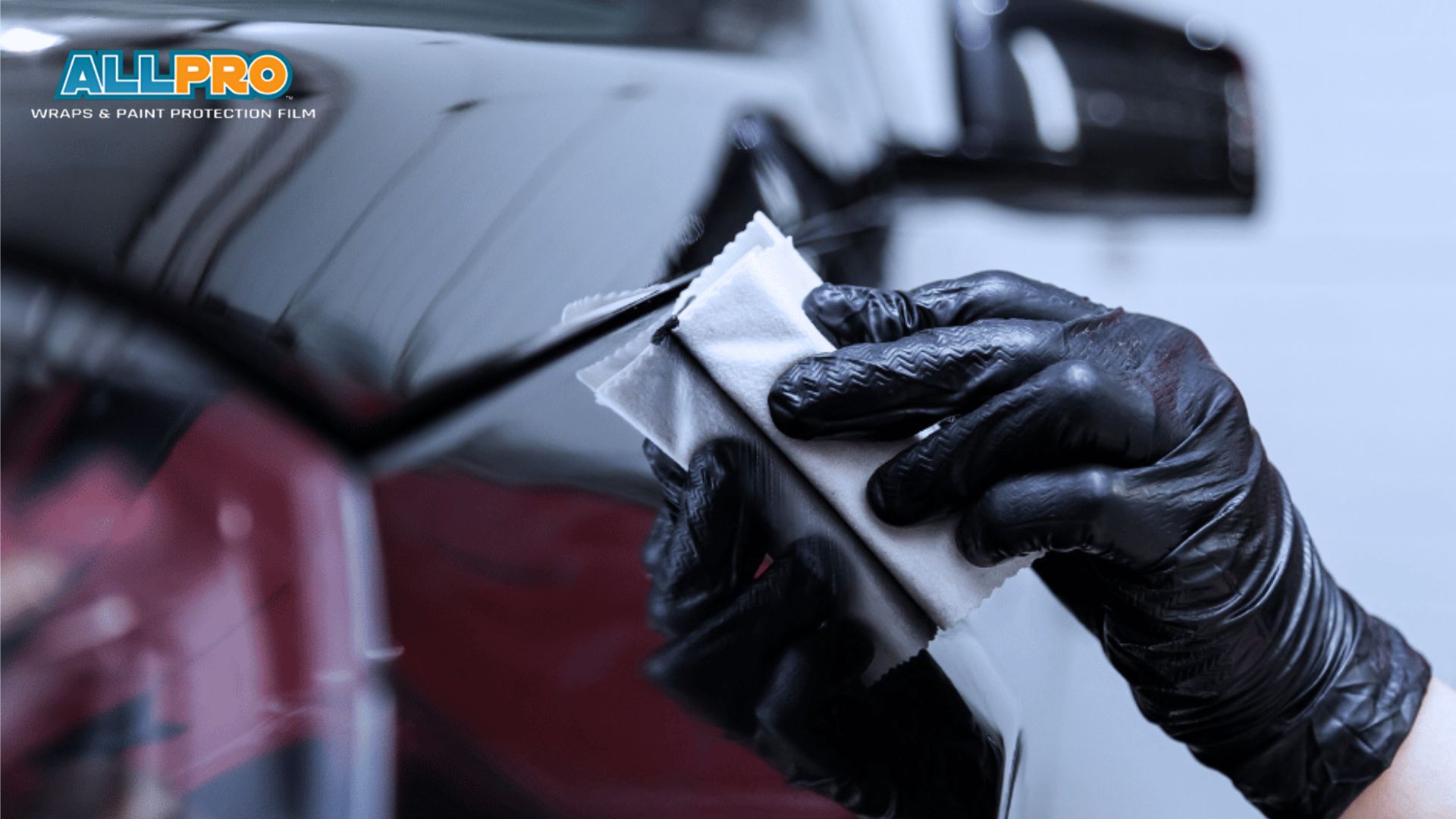 A technician in black gloves wipes the edge of a black vehicle panel near the “ALLPRO Wraps and Paint Protection Film” logo before ceramic coating. The close up shows precision surface preparation for a smooth glossy finish.