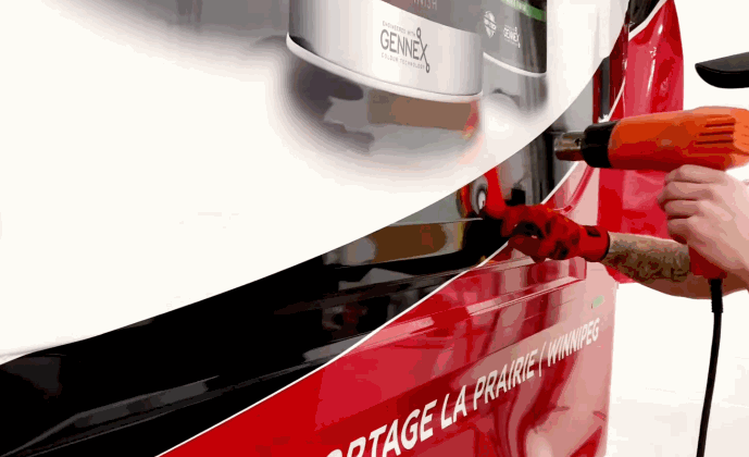 A technician uses a heat gun to apply commercial fleet graphics on the side of a red and white service van. Partial text on the vehicle reads “PORTAGE LA PRAIRIE WINNIPEG.”