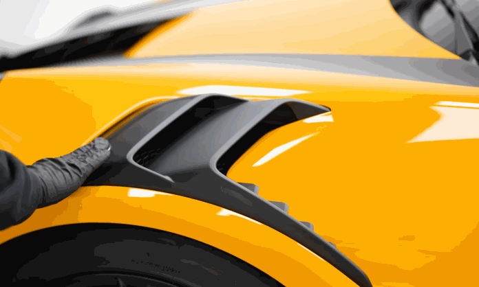 A technician in black gloves cleans a black side vent on a yellow sports car during car detailing. The close up shows careful work around the exterior trim.
