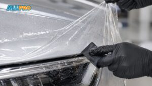 A technician uses a small squeegee to apply paint protection film to one section of a vehicle’s front bumper. The close up shows the clear film being smoothed over the painted surface for precise coverage.