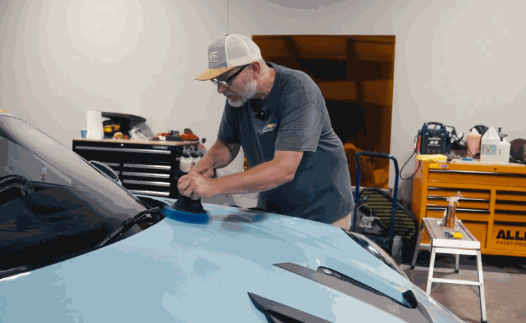 Technician polishes the hood of a light blue vehicle before ceramic coating in an auto detailing bay.