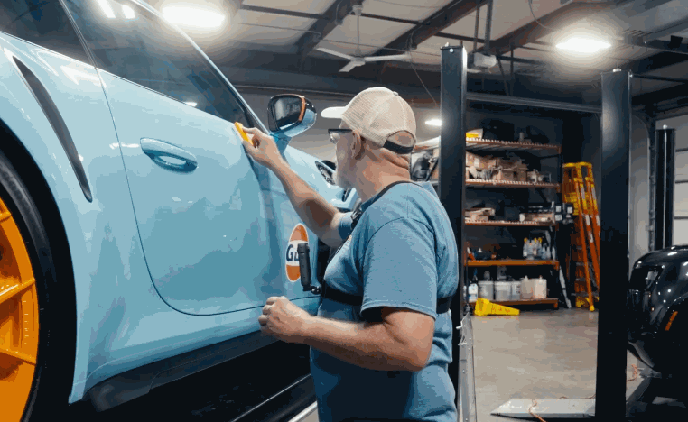 Technician applies ceramic coating around the door handle of a light blue vehicle in a detailing shop.