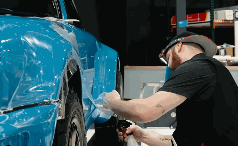Technician sprays and smooths paint protection film across the rear quarter panel of a blue car.