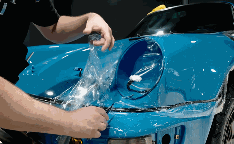 Technician applies paint protection film around the round headlight and front fender of a blue car.