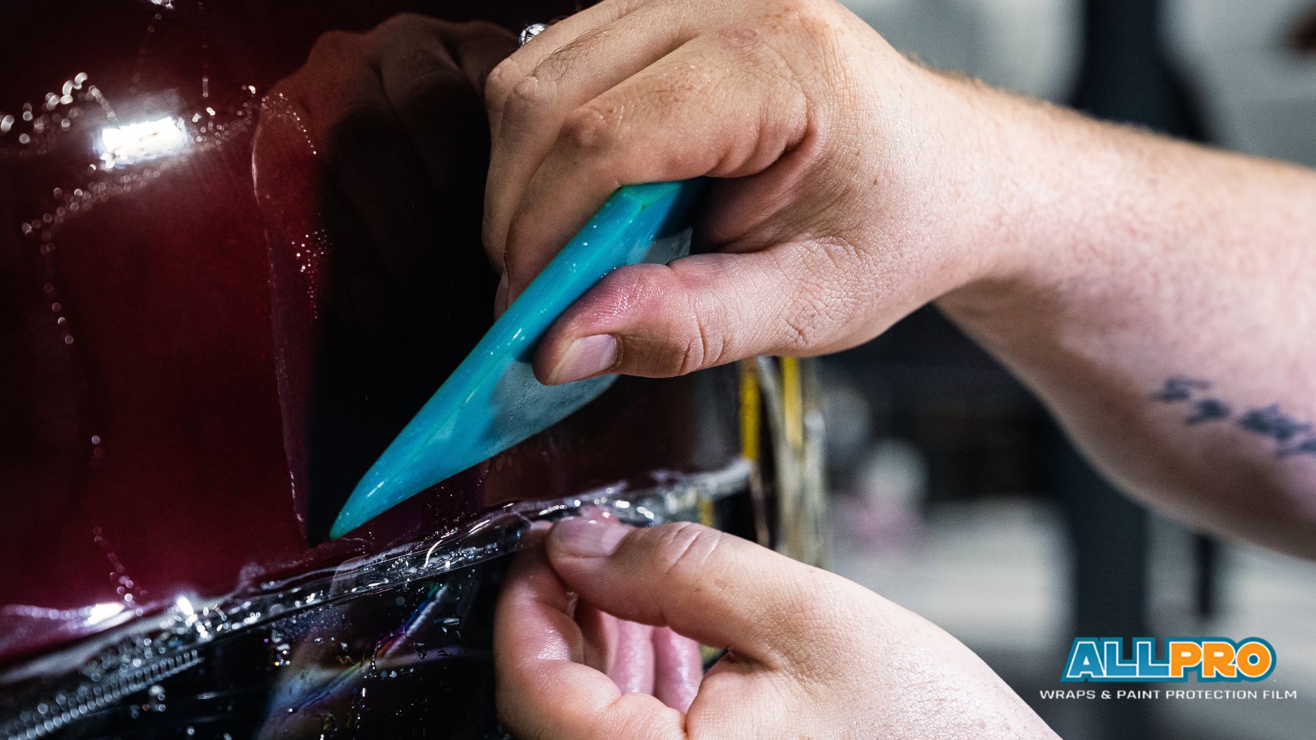 Technician uses a blue squeegee to tuck paint protection film along the headlight edge of a burgundy car.