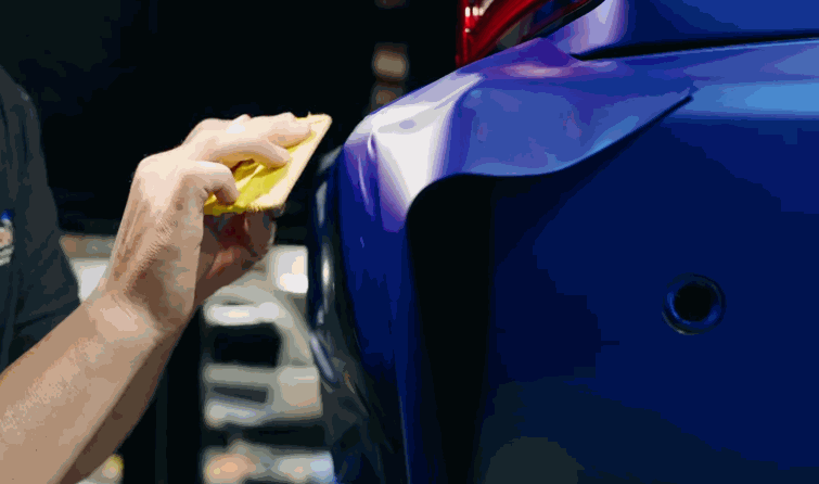 Installer smooths vinyl wrap around the rear bumper curve of a blue vehicle with a yellow squeegee.