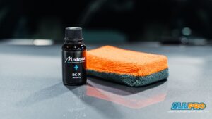 Bottle of ceramic coating sits beside orange and black microfiber towels on a vehicle surface.