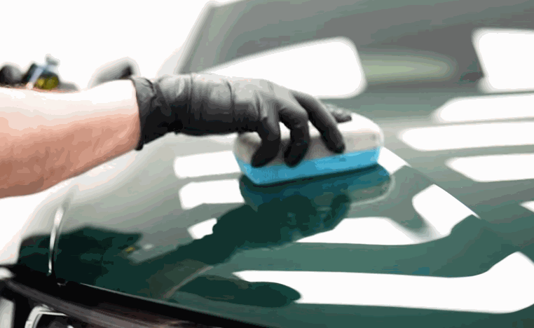 Detailer uses an applicator pad to spread ceramic coating across the glossy hood of a dark vehicle.