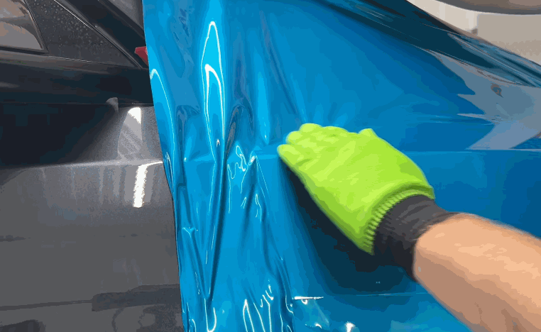 Installer wearing a bright green glove smooths glossy blue vinyl wrap across a recessed vehicle panel to remove wrinkles and help the material conform to the surface.